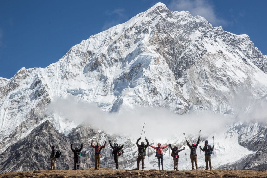 A group of hikers raising their arms for a photo with a snow covered mountain the background. They are hiking Everest Base Camp