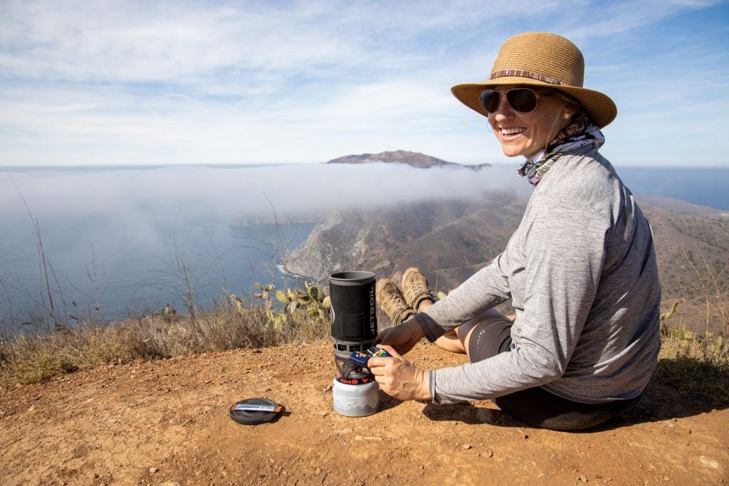 Kristen wearing Wallaroo Sun Hat while making coffee with jet boil on Catalina Island with ocean views in front of her
