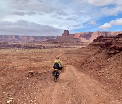 女性骑着满载的自行车在犹他州红色的土路上骑车，远处有红色的台地和悬崖