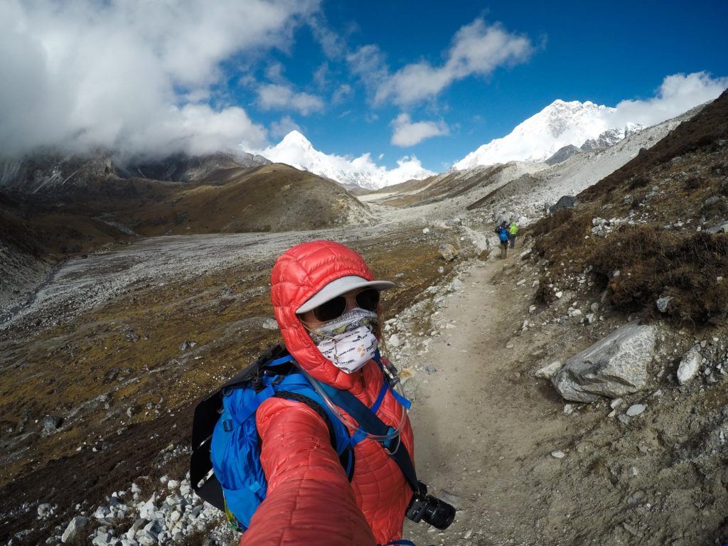 登山者在珠峰大本营小径上自拍，脸上覆盖着浅黄色，头上戴着夹克兜帽。背景是雪峰