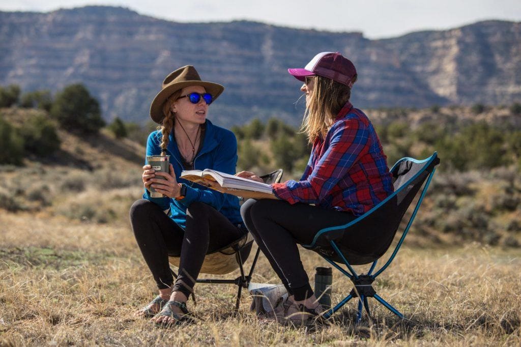 两个女人面对面坐在露营椅上聊天，背景是高耸的悬崖