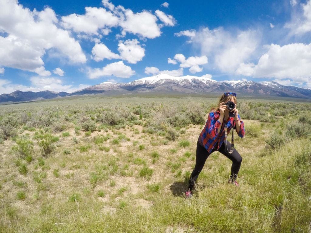 一名女子弯腰拿着相机，以山为背景拍照