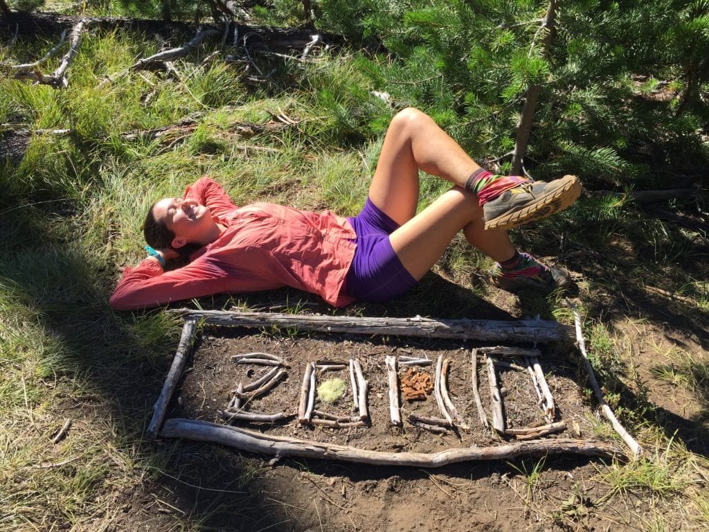 Woman hiker wearing pink jacket and purple shorts lying on back behind sign made of wood and sticks reading '2000'. This marks mile 2,000 of her Pacific Crest Trail thru-hike.