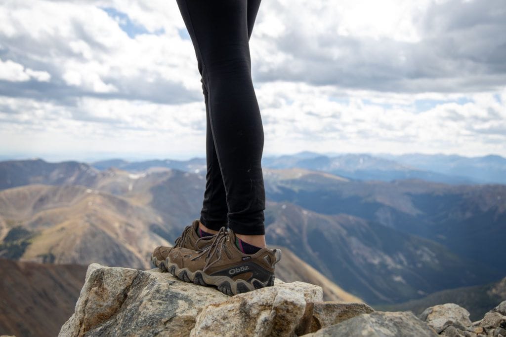 Close-up photo of woman wearing low-top Oboz hiking boots standing on rock ledge with mountain ranges in background