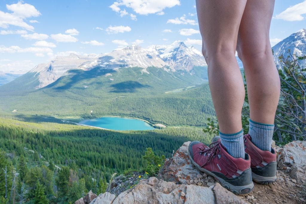 A woman stands on a summit looking over  a blue lake and lots of alpine trees. She's wearing red Oboz hiking boots