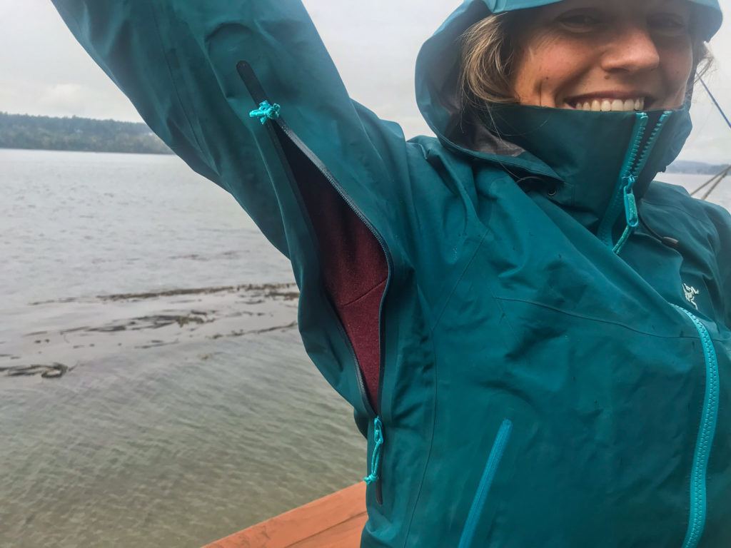 woman hiking in the rain next to a lake with her arm raised, exposing an open zipper on her rain jacket in the armpit