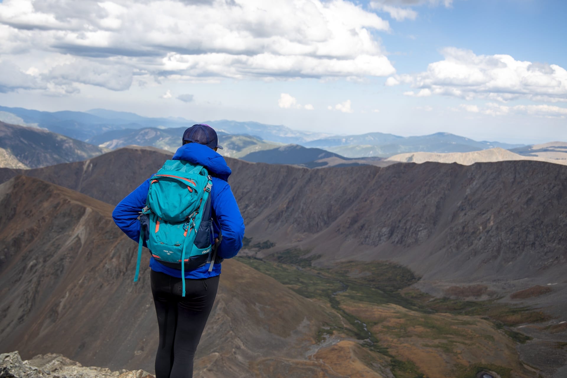 Female hiker standing on top of mountain looking out over nearby mountain ranges. She's wearing a blue jacket and blue hiking day pack.