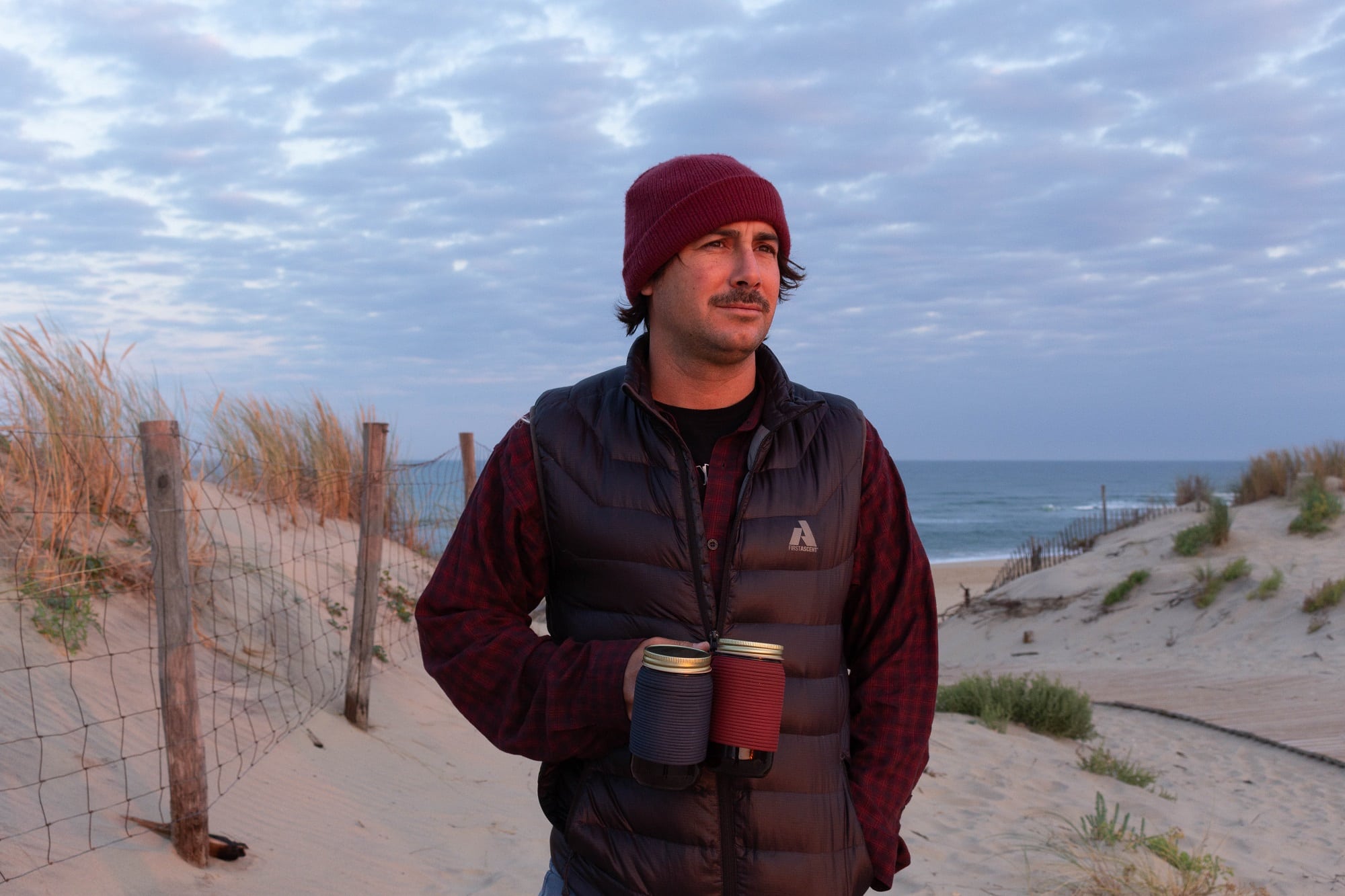 日落时分，穿着保暖衣服的男子在海滩上拿着两个绝缘马克杯