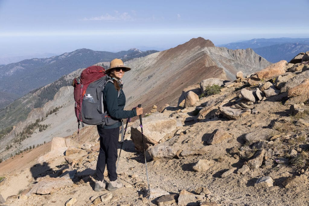 在红杉国家公园，一名妇女穿着登山硬装Dynama踝裤，背着红色背包站在岩石山脊上微笑
