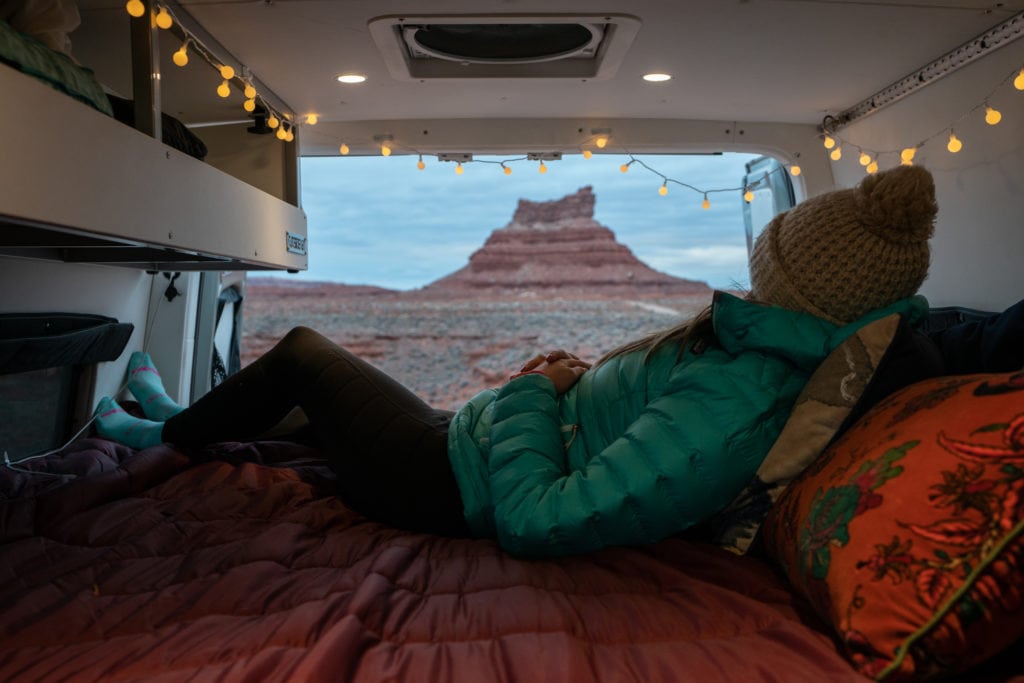 一个女人躺在她露营车的床上，后门开着，上面盖着一条Rumpl毯子