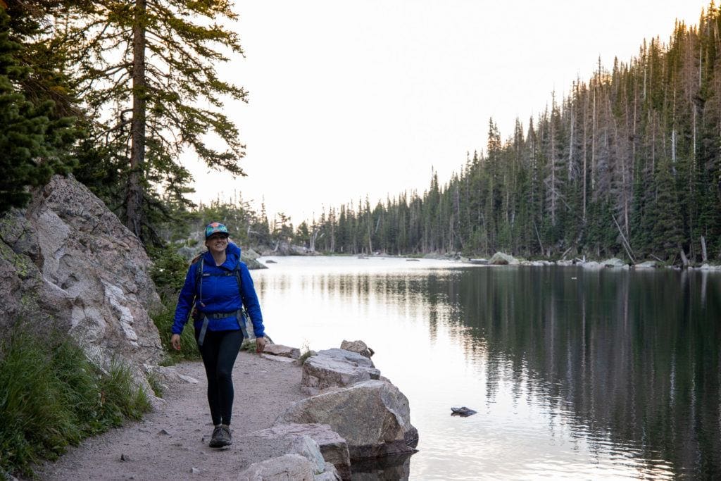 Woman hiking on a trail alongside a lake during sunset with a forested ridgeline in the background