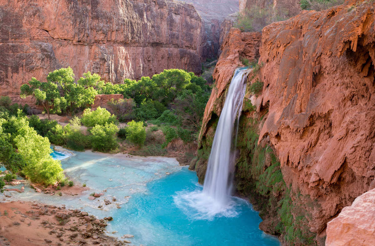 View of Havasu Falls from above with waterfall cascading into turquoise blue water surrounded by red rock cliffs