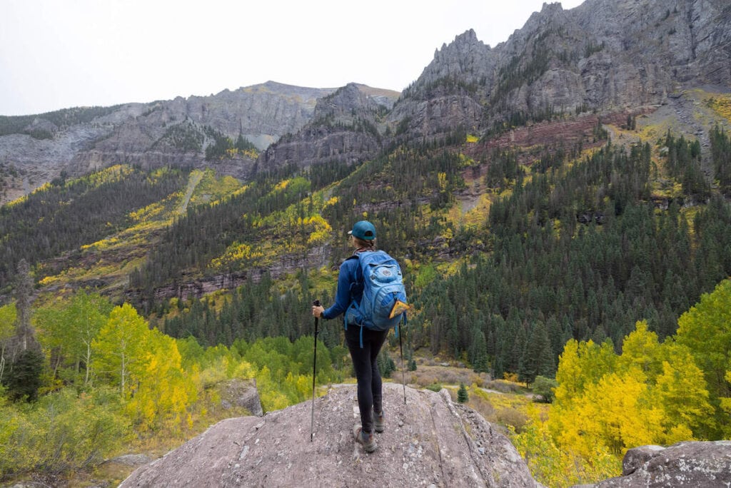 克里斯汀站在科罗拉多州的岩壁上，背着蓝色的鱼鹰背包，手拿登山杖，眺望山脉