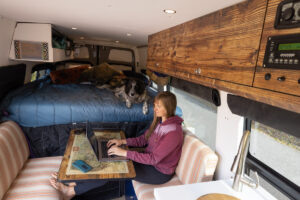 一名妇女坐在拉贡桌子前，在她的露营车里用笔记本电脑工作，背景是一张固定的床