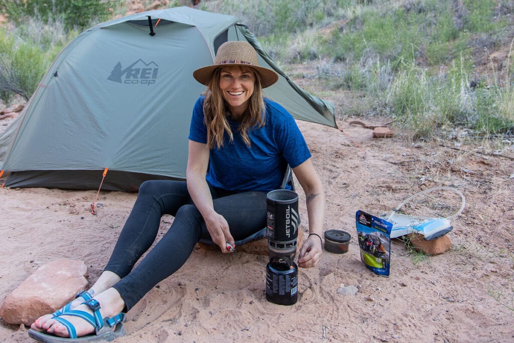 Woman sitting on the ground in front of a backpacking tent cooking on a backpacking stove