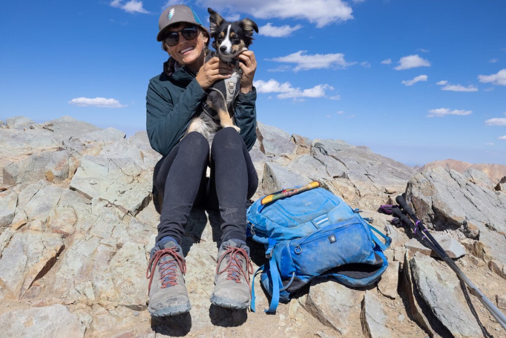 克里斯汀坐在岩石堆上，穿着Oboz Sypes登山鞋，在徒步旅行的顶峰，抱着小狗和背包在她身边
