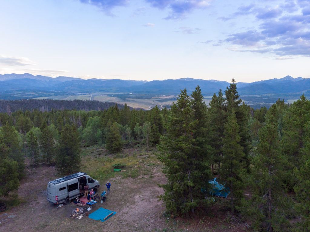 Van parked in dispersed campsite in Colorado with tent set up in trees and campfire going with mountain range in distance