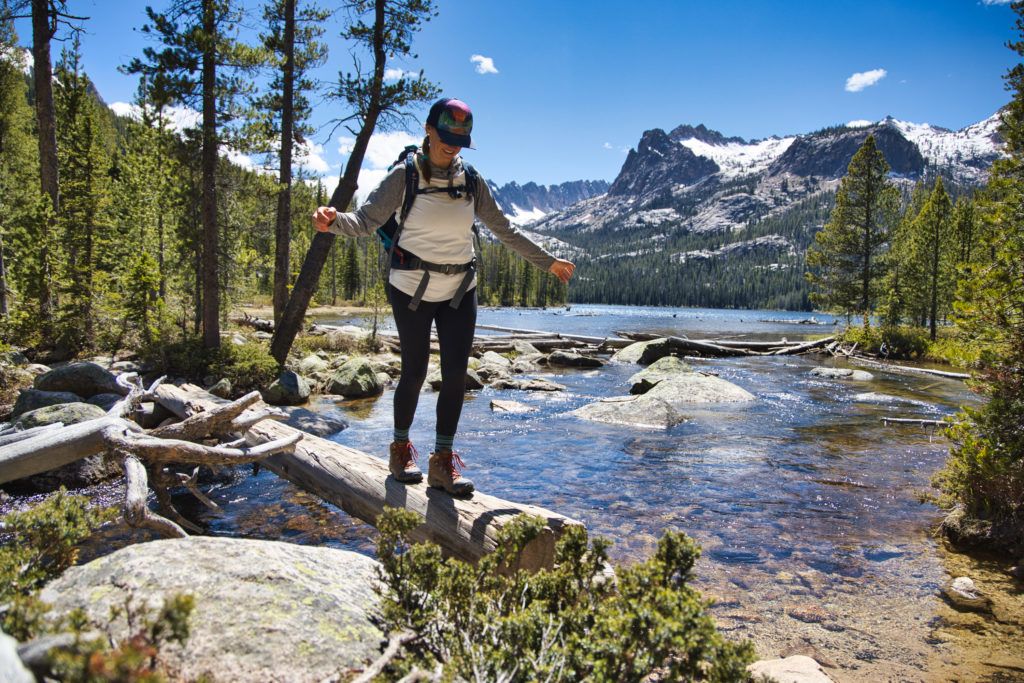A woman balances on a log while on a hike. There is a lake behind her.