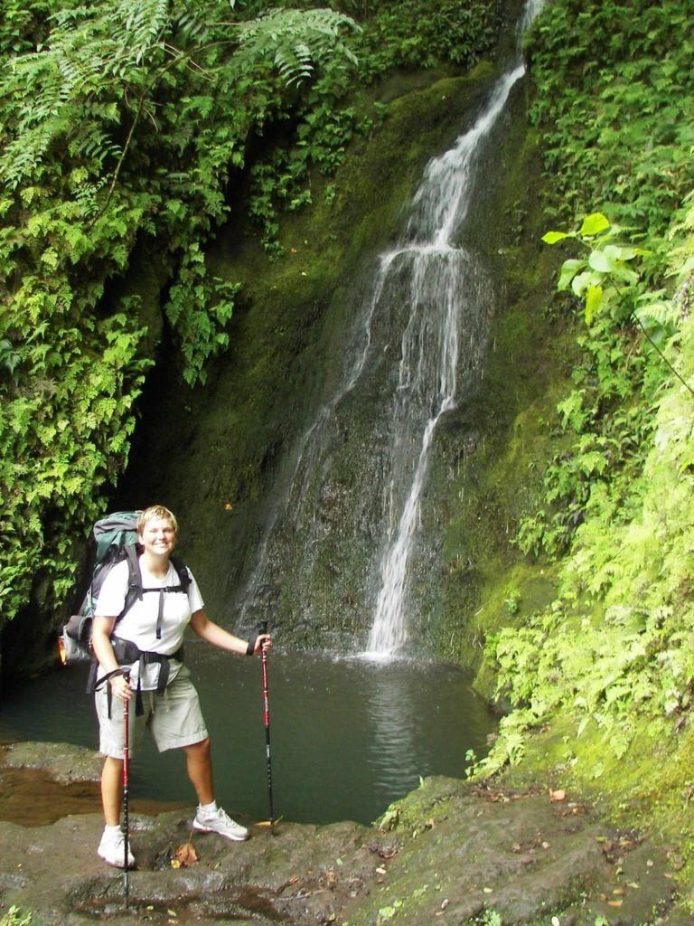 A woman stands next to a waterfall in Hawaii with a hiking backpack on and trekking poles