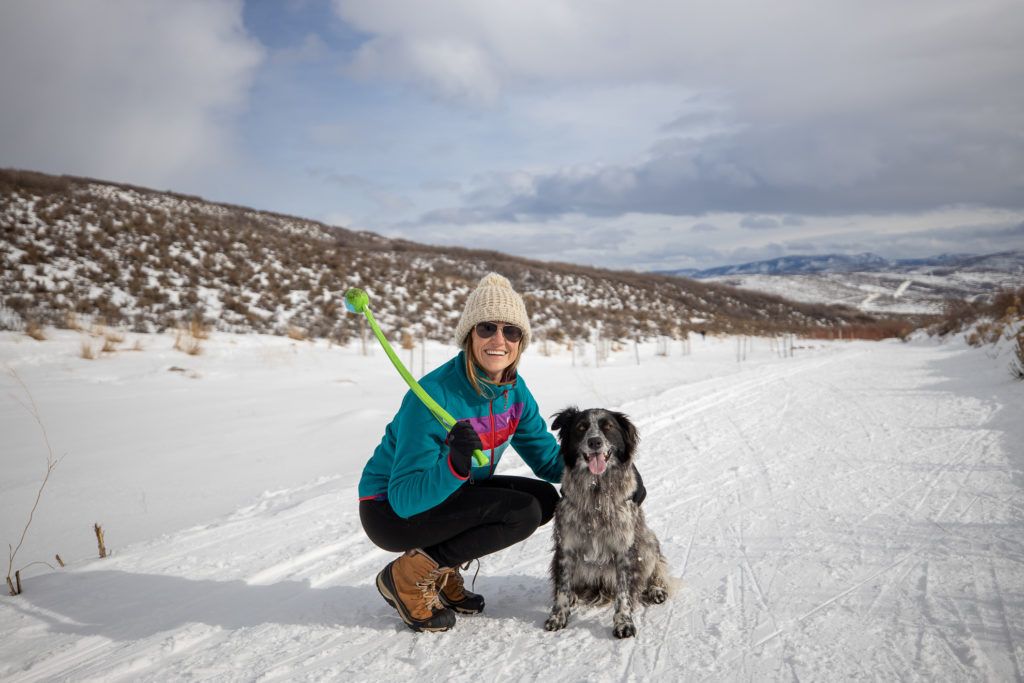 克里斯汀蹲在冬天徒步旅行的小路上，一只手拿着塑料球，在狗旁边摆姿势拍照