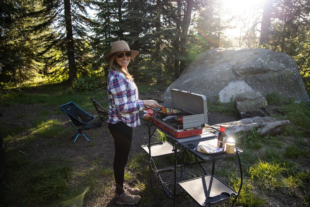 A woman cooks while camping using the GCI Outdoor Slim Fold Cook Station