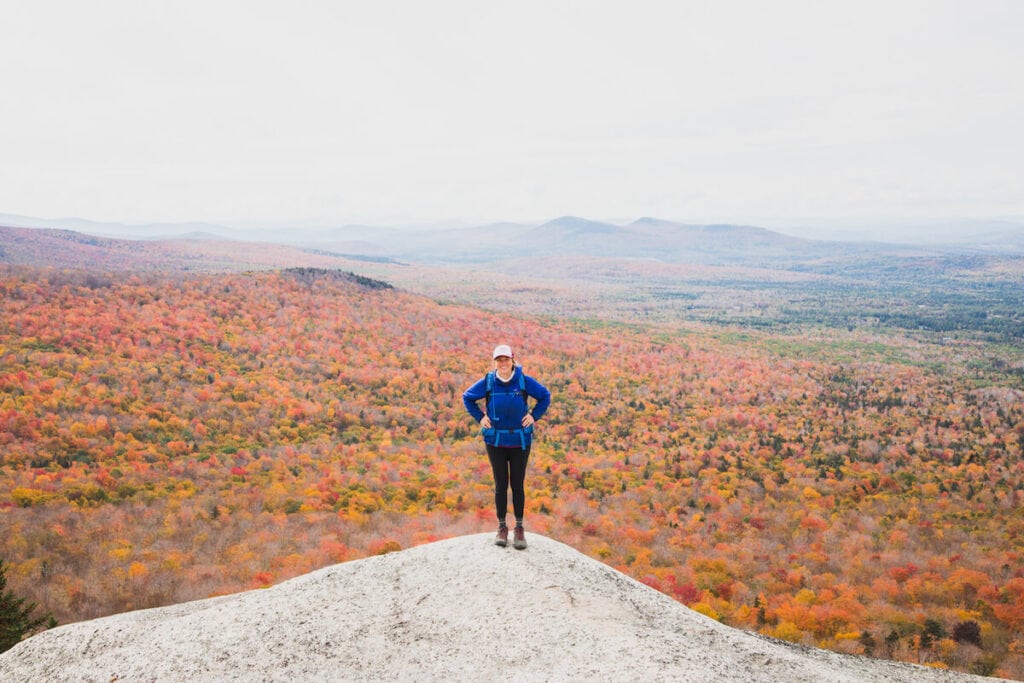 Woman standing on rock ledge overlock facing camera with expansive fall foliage forest and colors behind her