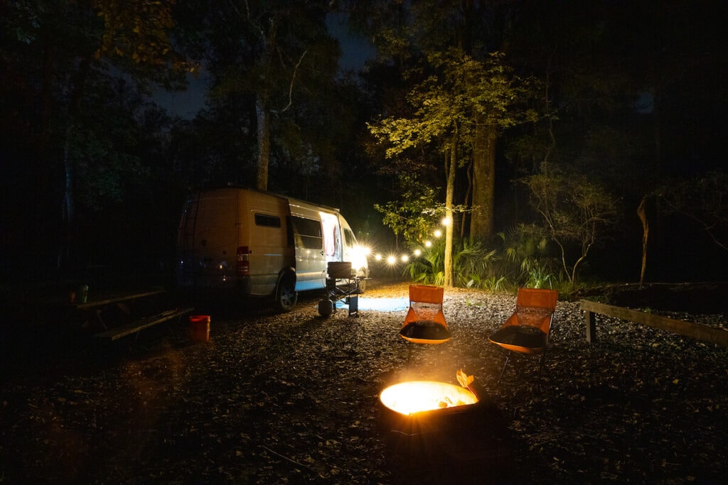 Sprinter Camper Van parked at a campsite at night with two camp chairs set up next to a campfire with string lights hanging for a nice ambiance