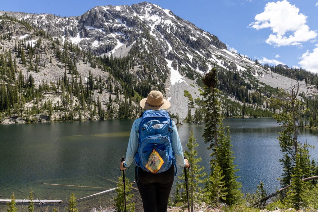 Woman standing at edge of high alpine lake with peak dusted with snow. She is wearing day hiking pack and carrying trekking poles
