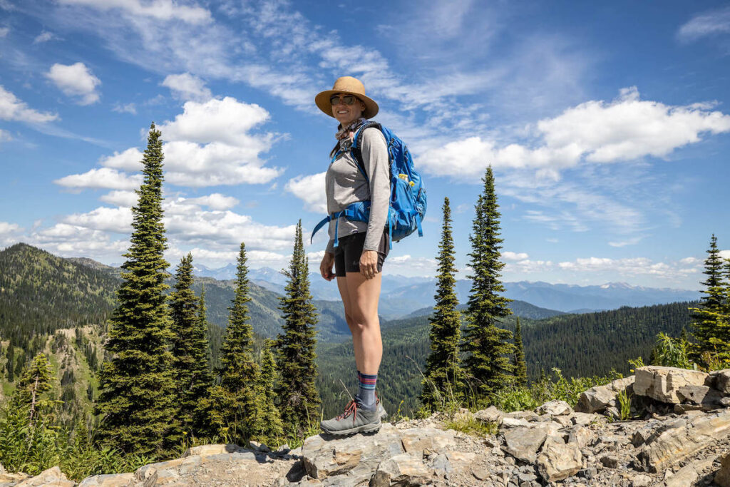 Kristen wearing mid-ankle Oboz Sypes hiking boots on rocky trail in Montana. Pine trees and mountain ranges in the background.