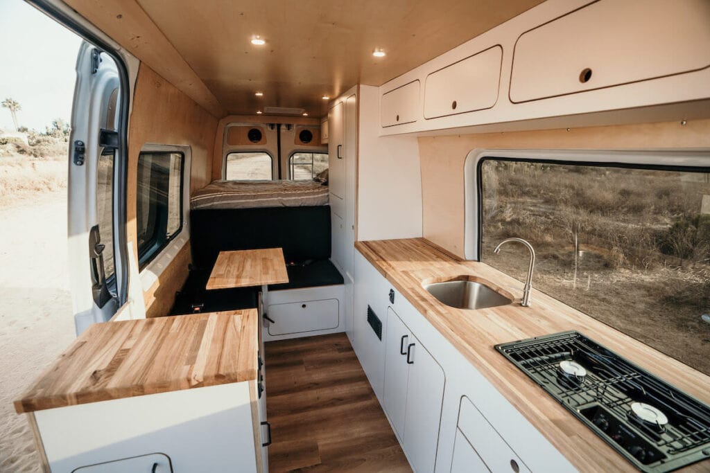 Beautiful interior of one of VanCraft's rental campervans showing wood floors and countertops, a sink, stove, bed, and more