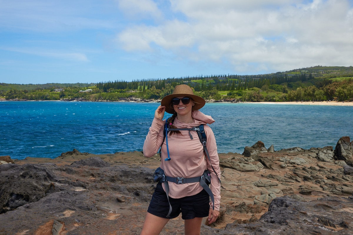一名女子在毛伊岛的海边徒步旅行，她穿着远足短裤、长袖防晒衬衫、帽子和远足背包