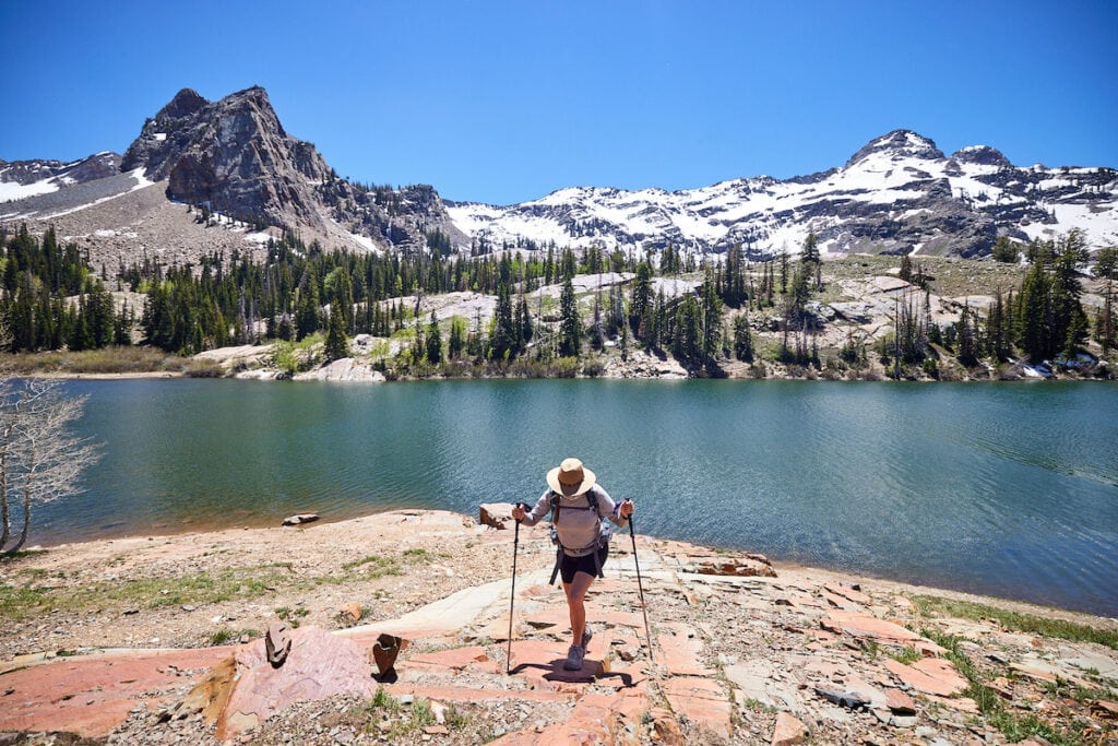 Woman hiking uphill on a rocky trail with a lake and snow capped mountains in the background