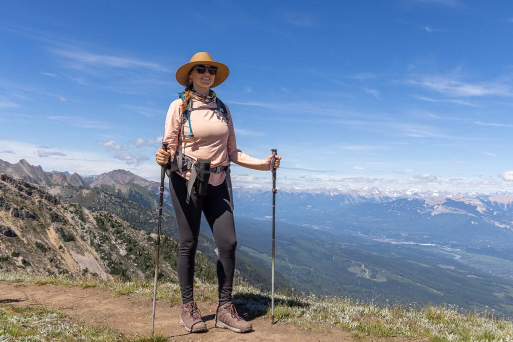 Woman hiking on a ridgeline in BC wearing leggings, a long sleeve shirt for sun protection, a hat, a hiking backpack, and holding trekking poles