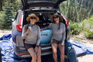 Two women sitting in rear of car packed with camping gear and Artic cooler