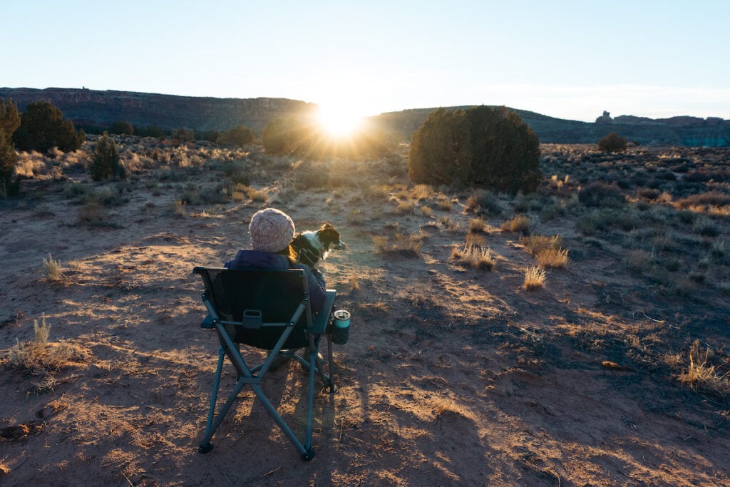 犹他州摩押，一名妇女坐在雪人露营椅上，她的腿上躺着一只狗，太阳在他们面前落下