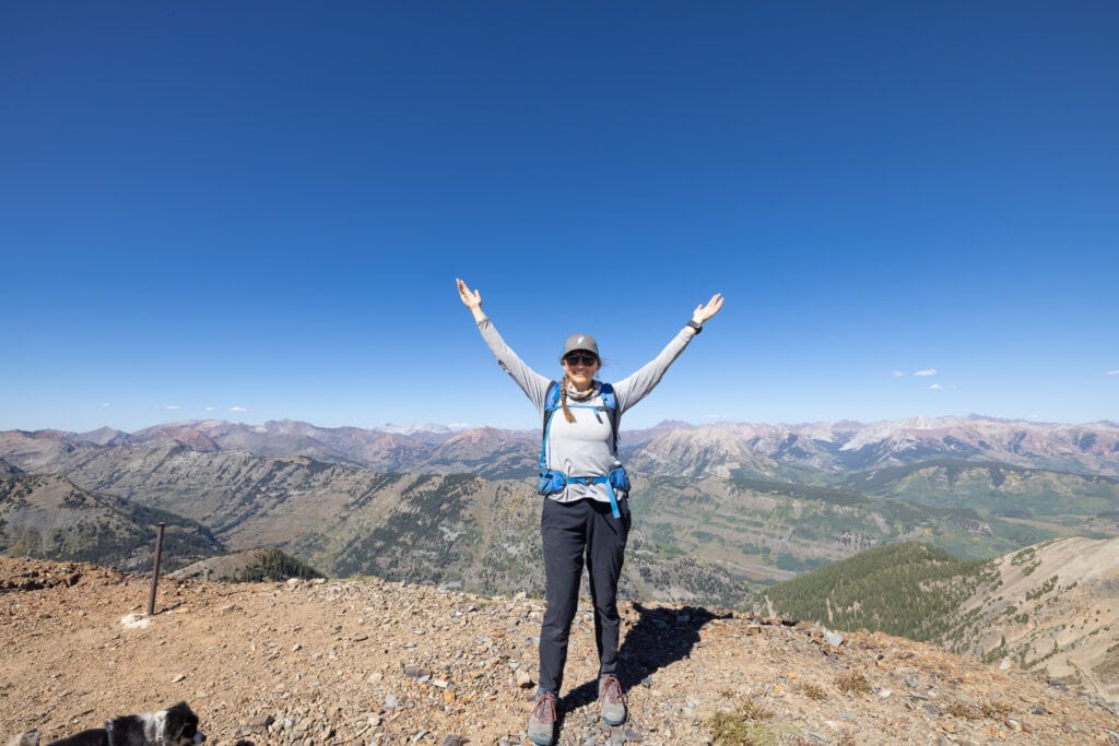 Kristen standing at lookout on trail in Colorado with both arms raised above her head