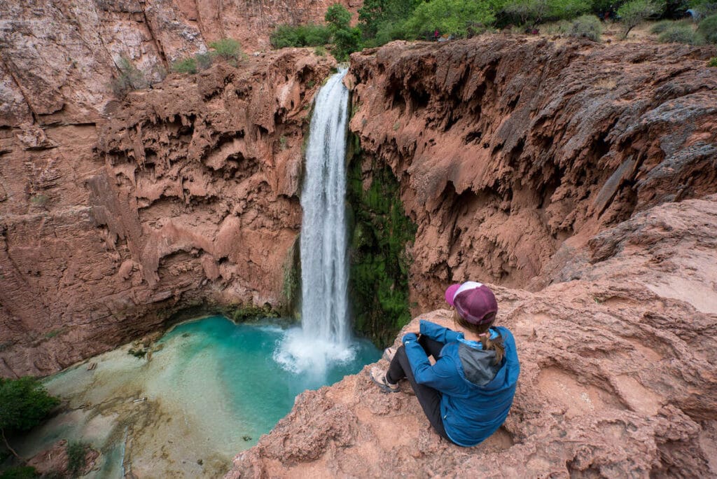 Woman sitting on cliff edge overlooking Havasu Falls and turquoise blue pool at base