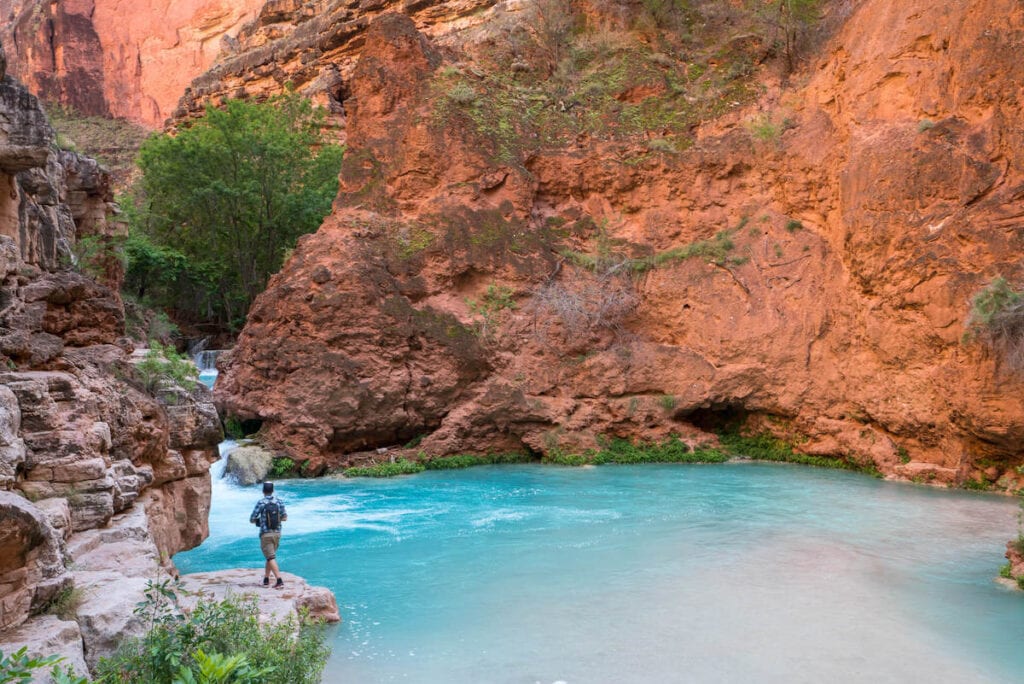 妈n standing on ledge above turquoise blue waters surrounded by red rock cliffs in Havasu Canyon in Arizona