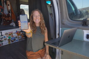 一个女人坐在她的面包车门口，手里拿着一部屏幕上有可视无线标志的手机，旁边的桌子上放着一台笔记本电脑