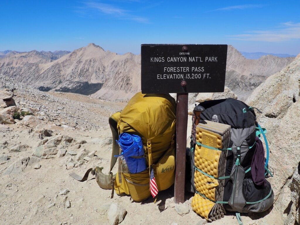 两个背包坐在JMT上写着“进入国王峡谷国家公园”的标志旁边
