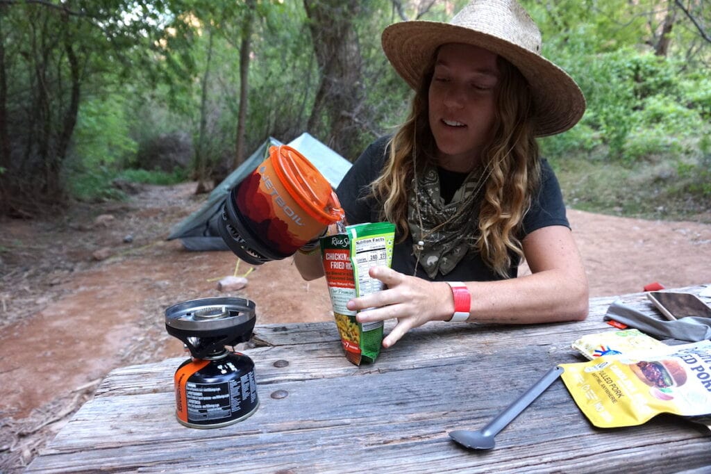 a woman sits at a picnic table pouring hot water from a Jetboil Mini Mo into a Knorrs Rice pouch