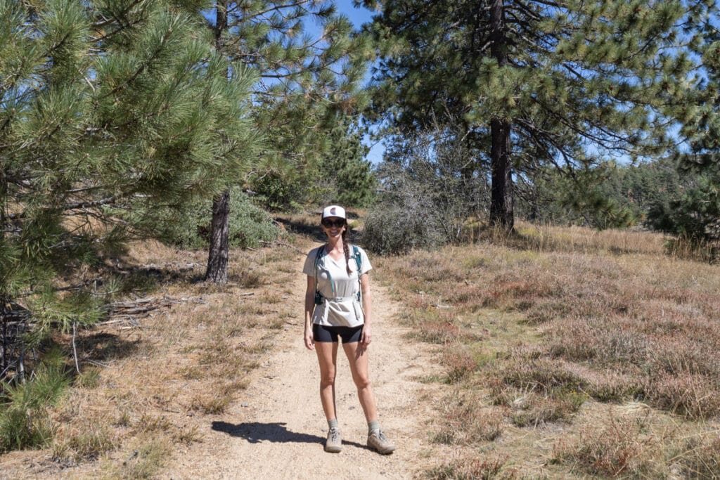 Woman smiles at the camera standing on a hiking trail. She is wearing a white REI Co-op Sahara T Shirt