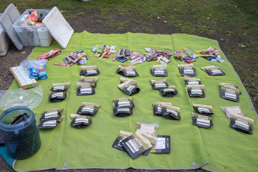 A green tarp is laid out on the ground with a BearVault 500 bear canister in the bottom corner. There are Outdoor Herbivore backpacking meals and various snacks organized on the mat.