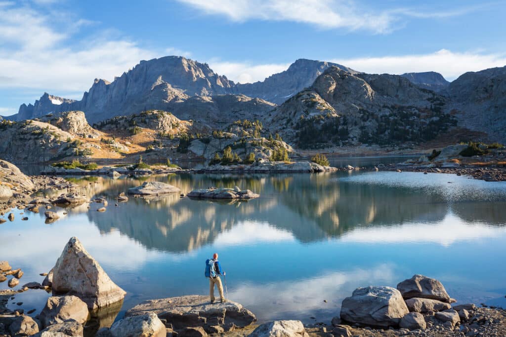 A man stands on a rocky ledge next to a lake. He's backpacking the Cirque of the Towers in the Wind River Range in Wyoming