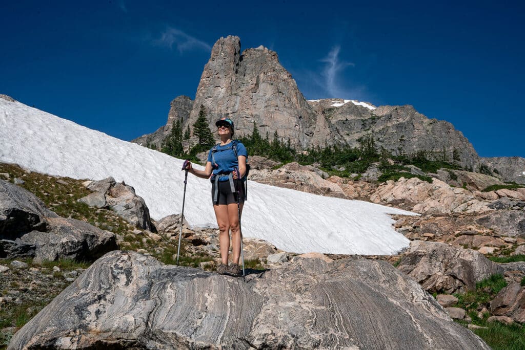 Woman hiker holding trekking poles standing on a large rock with rocky mountains and snow in the background