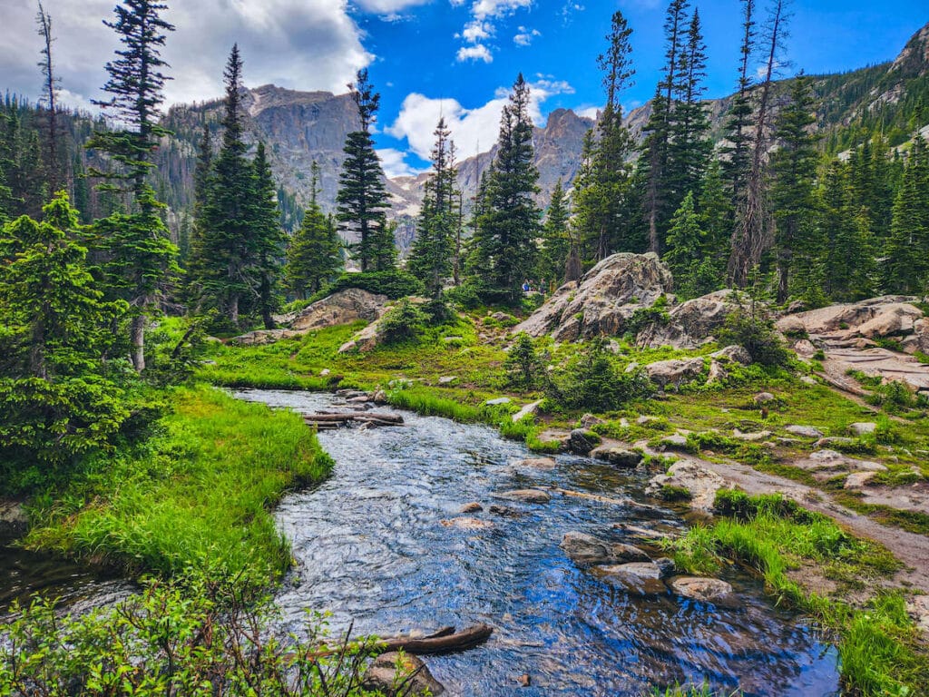 Alpine river leading from Dream Lake in Rocky Mountain National Park surrounded by vibrant green grass and trees and tall, rocky mountains in the distance