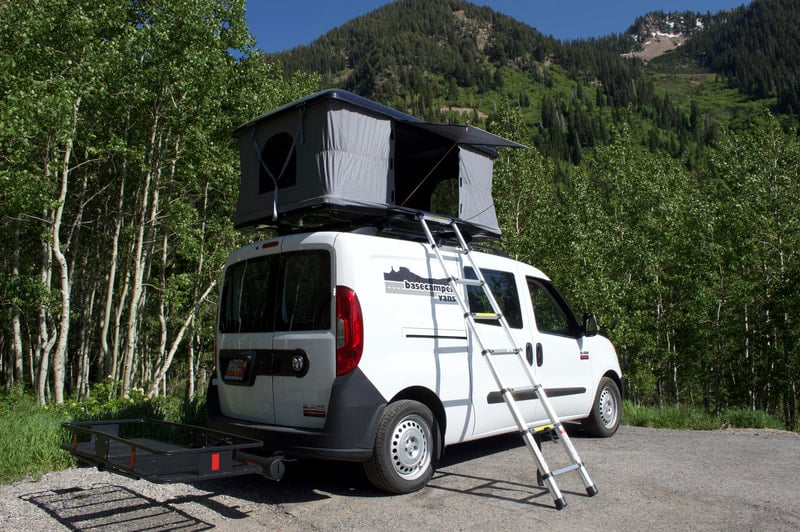 White Dodge Promaster City campervan with a rooftop tent set up at a campsite in front of trees