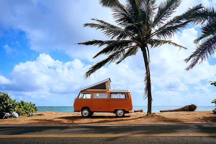 A bright orange classic Volkswagon campervan parked on the beach in Hawaii under a palm tree available for rent via Hawaii Surf Camper Vans