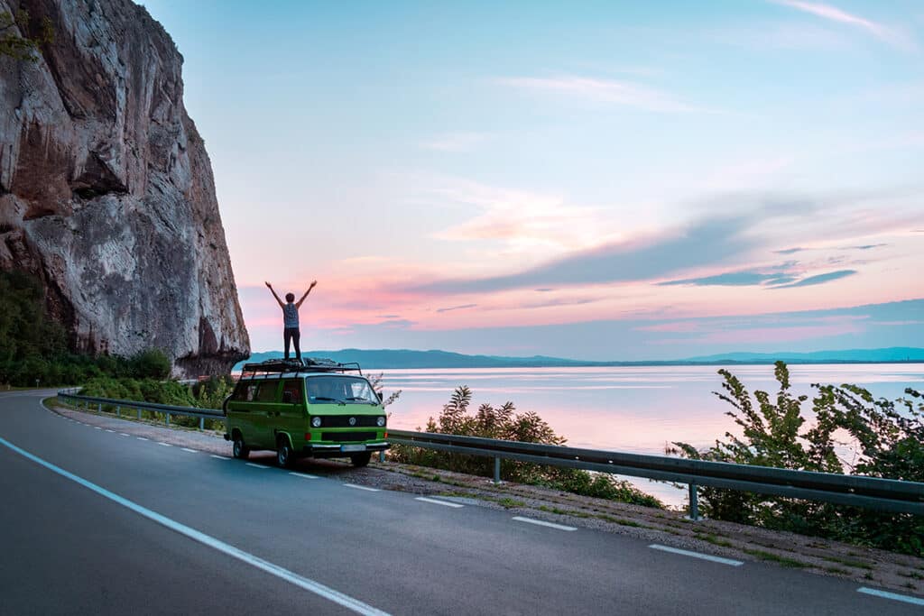 女人站在一个绿色的大众Vanagon露营者van parked on the side of the road alongside the ocean at sunset